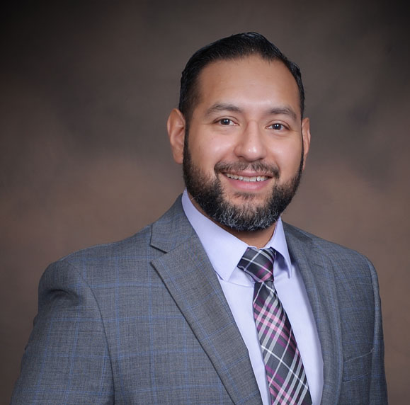 Mike Negrete in a formal gray suit with a plaid tie, standing against a neutral backdrop professionally.