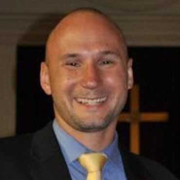 Christopher Shilling, wearing a black suit, light blue shirt, and yellow tie, stands before a backdrop with a subtle cross.