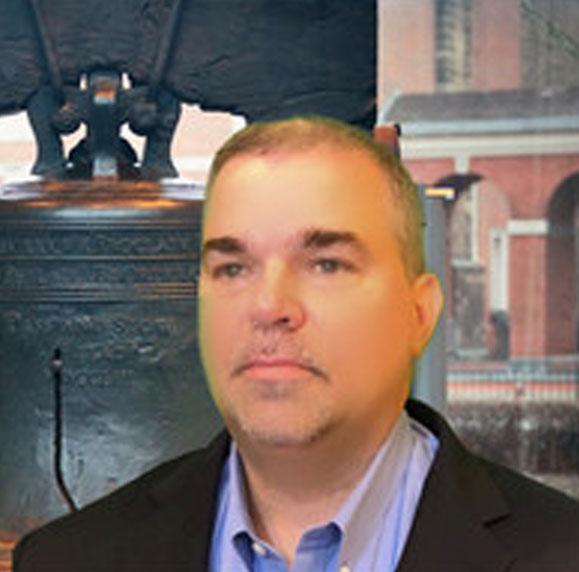 Scoot Battaglini, wearing a black suit, light blue shirt, and yellow tie, stands before a backdrop with a subtle cross.